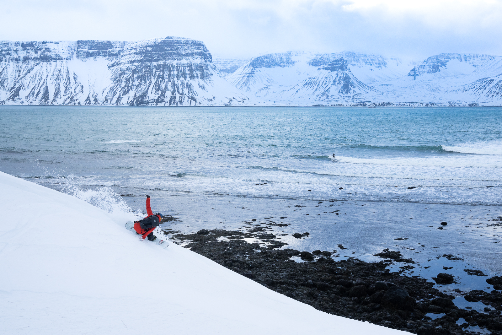 Elena Hight snowboards while Peter Devries surfs in the background. Westfjords, Iceland.